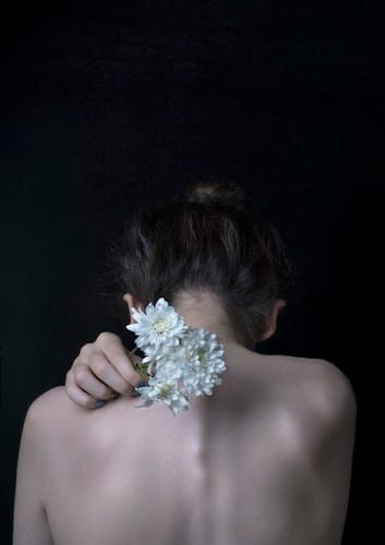 Dos nu d’une femme qui tient des fleurs blanches sur son épaule.