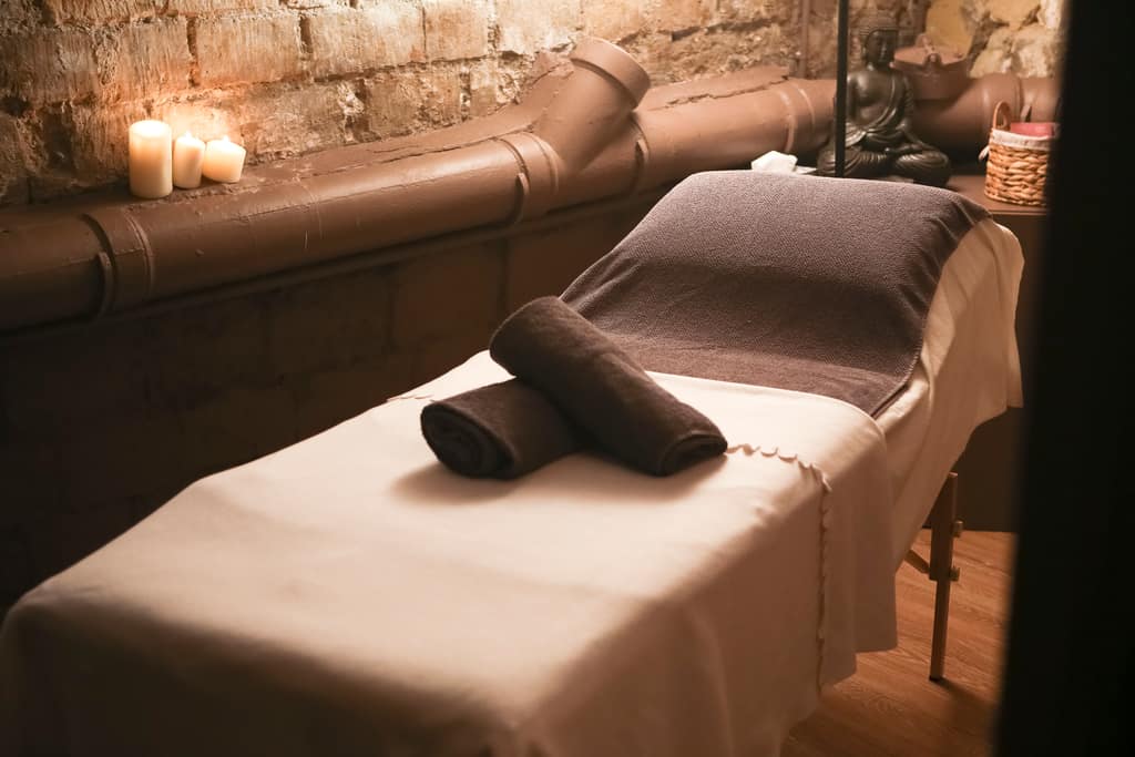 Salle de soins à la lumière des bougies, table couverte d’un drap clair et de serviettes roulées.