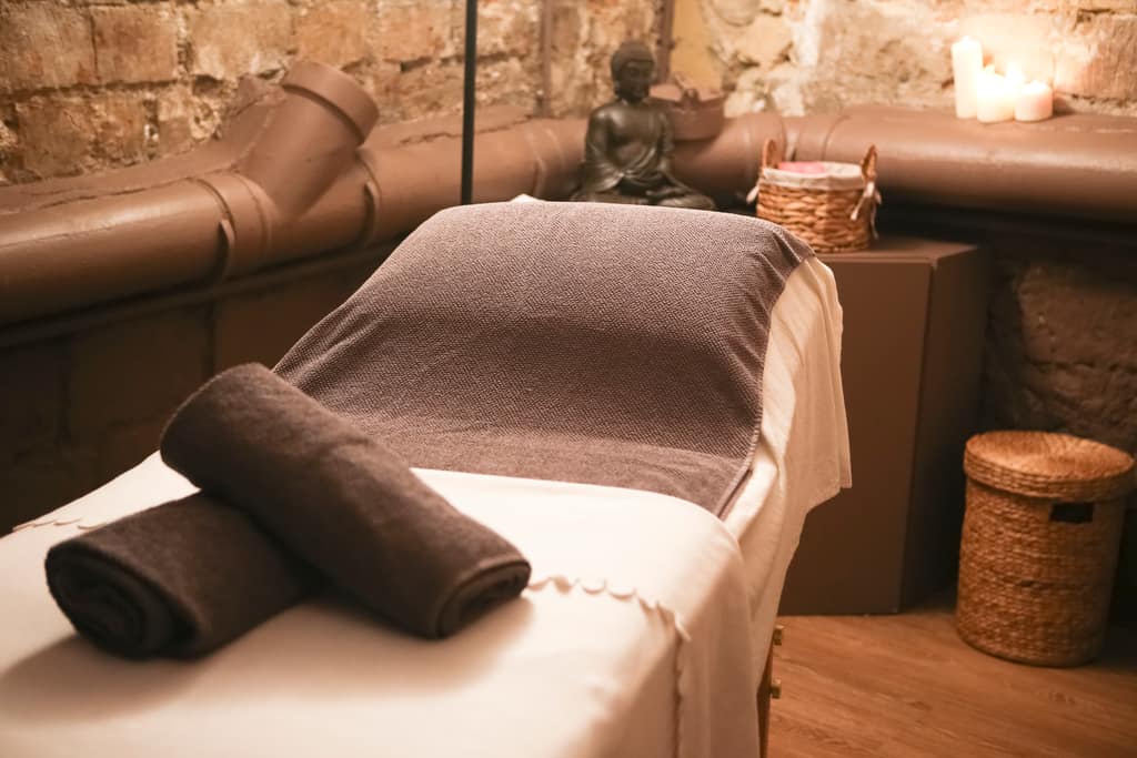 Table de massage préparée avec des serviettes roulées, devant un mur de briques éclairé de bougies.